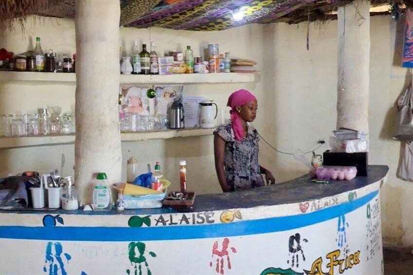 A bar counter with a woman standing behind it, shelves with bottles and glasses, and a colorful mural on the wall. 2