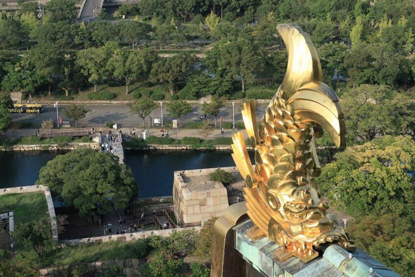 Una scultura dorata a forma di pesce su un ponte, con un fiume e vegetazione sullo sfondo. 2
