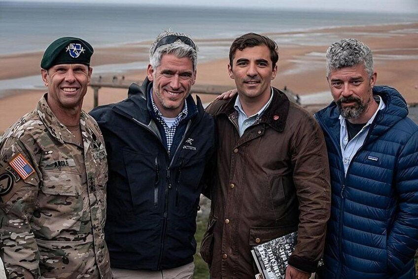 Vier mensen staan op het strand, met een wateroppervlak op de achtergrond. 5