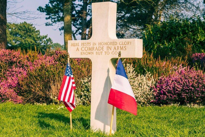 Een kruis met een inscriptie, omringd door gras en bloemen, met een Amerikaanse vlag en een Franse vlag. 3