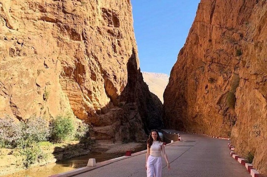 A person standing on a road with red and white curbs, surrounded by red rock cliffs and a small body of water. 3