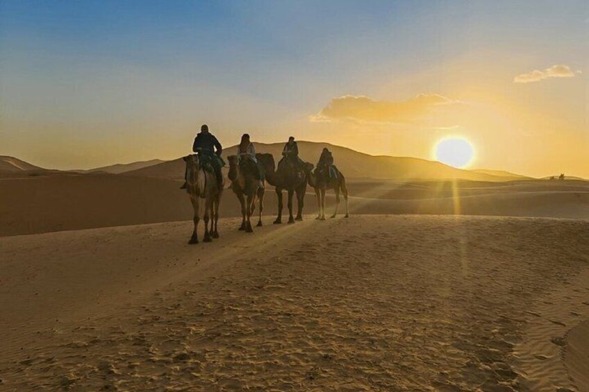 A group of people riding camels on a desert trail during sunset. 3