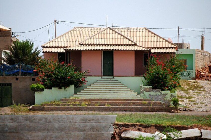 Una casa con techo de tejas, una puerta verde y una escalera que conduce a la entrada. Hay dos macizos de flores con flores rojas a ambos lados de la casa. 2