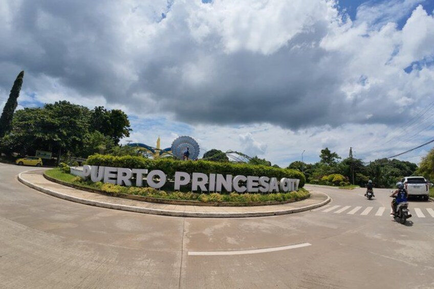 Una rotonda con un cartello che indica 'PUERTO PRINCESA CITY' e una ruota panoramica sullo sfondo. 3