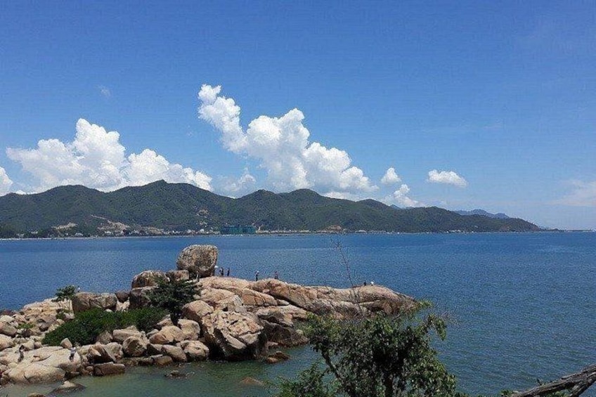 Una costa rocosa con agua azul cristalina, montañas a lo lejos y algunas personas pescando. 2