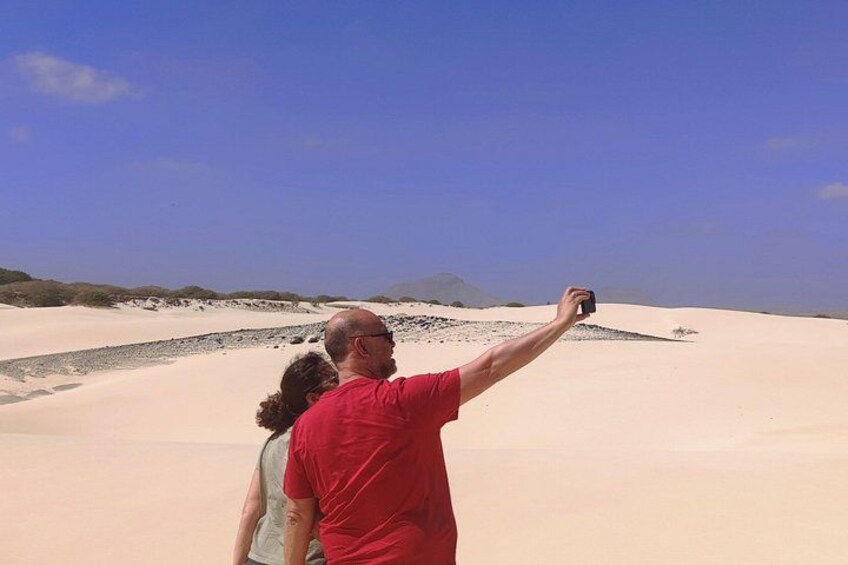 Eine Person macht ein Selfie mit einer Kamera, im Hintergrund eine weitere Person und Sanddünen. 4
