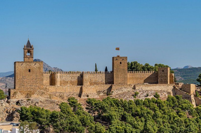 A historic stone fortress with towers and battlements, surrounded by greenery and mountains. 2