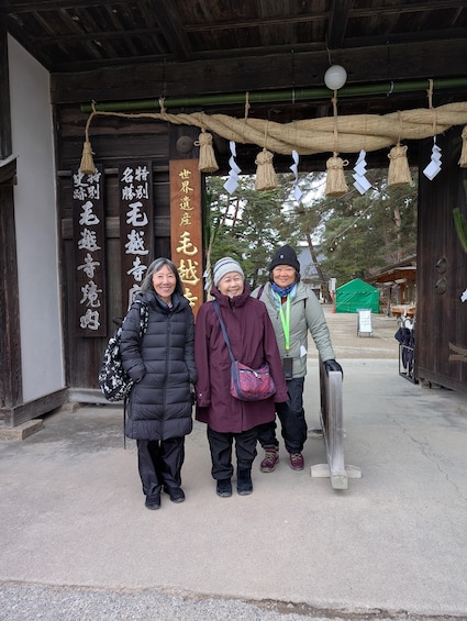 Three people standing in front of a traditional wooden structure with hanging decorations and signage. 2