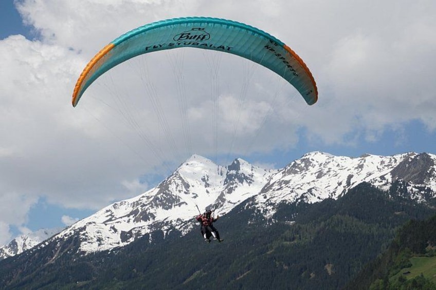 Een persoon paraglide over een bergketen met sneeuw, met een blauwe en oranje vlieger. 4
