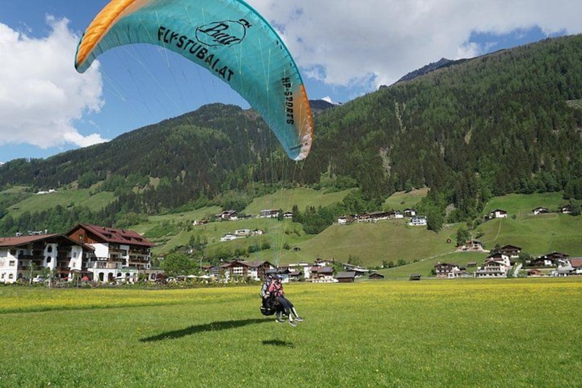 Een persoon paraglidet met een banner, een grasveld en een bergachtig landschap met gebouwen op de achtergrond. 3