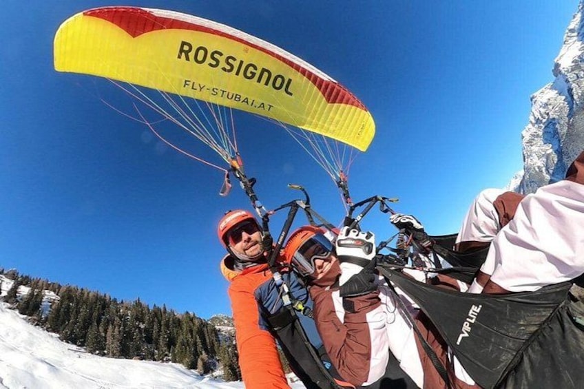 Een persoon paraglidet met een gele en rode parachute met de tekst 'ROSSIGNOL' en 'FLY-STUBAIAT' voor een besneeuwde berg. 5
