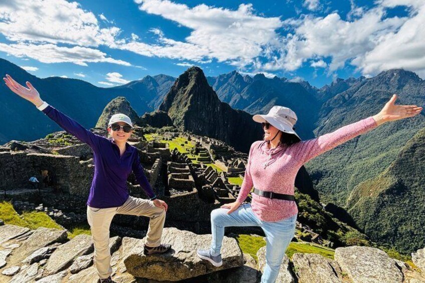 Two people standing on a rocky outcrop with arms outstretched, overlooking Machu Picchu ruins and surrounding mountains. 4