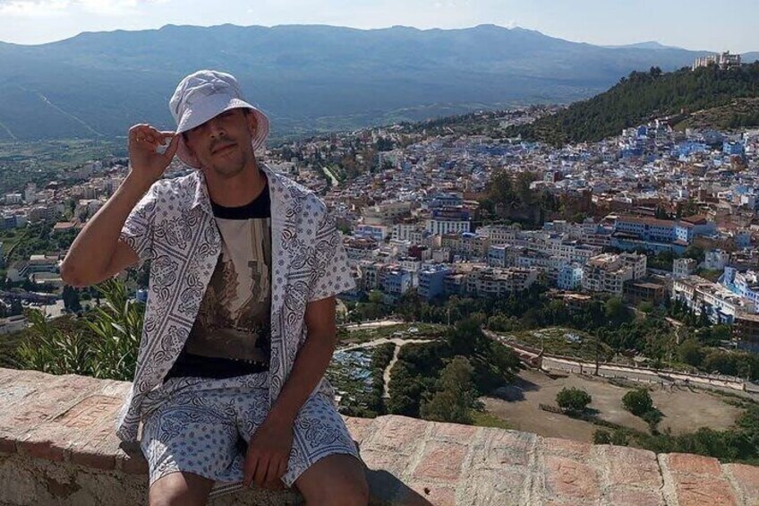 A person sitting on a ledge overlooking a town with blue buildings and a mountainous backdrop. 3