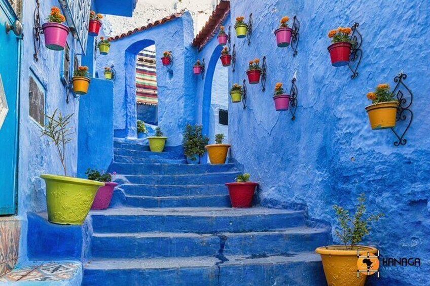 A narrow blue-painted alley with colorful hanging pots and steps. 4
