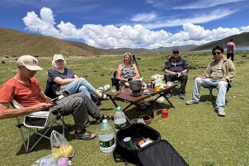 A group of people sitting on chairs and camping tables in a grassy field with mountains in the background. 3