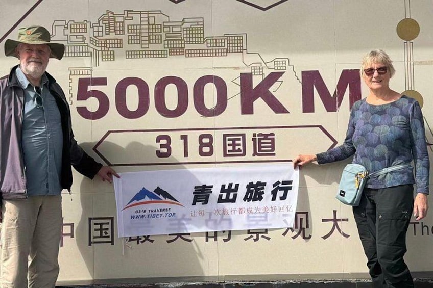 Two people standing in front of a sign with '5000KM' and '318 National Highway' text. 5
