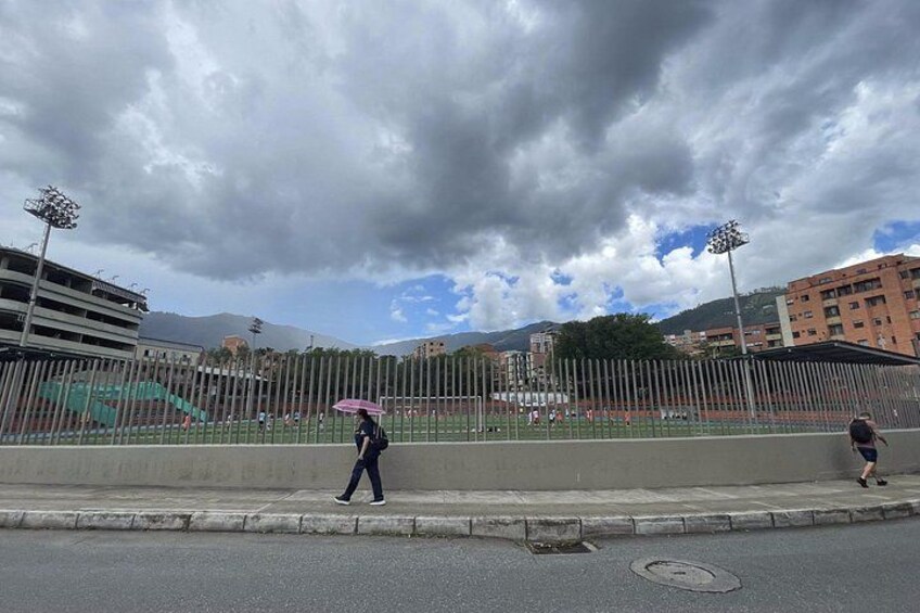 Un peatón cruzando una calle bajo un cielo nublado, con edificios y un campo deportivo al fondo. 3