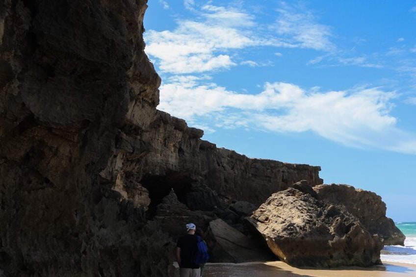 A rocky cliff with a cave entrance, a person standing near the cave, and a beach in the background. 2