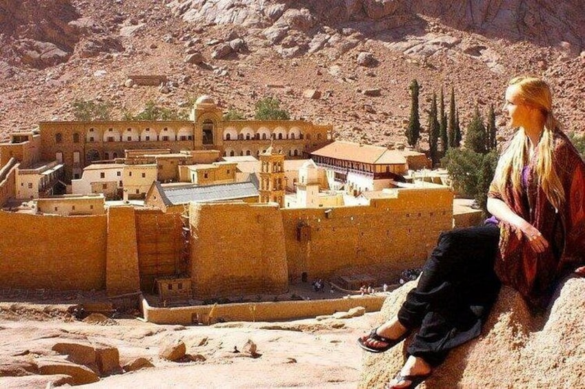 A desert landscape with a large, ancient walled city and a person sitting on a sand dune. 1
