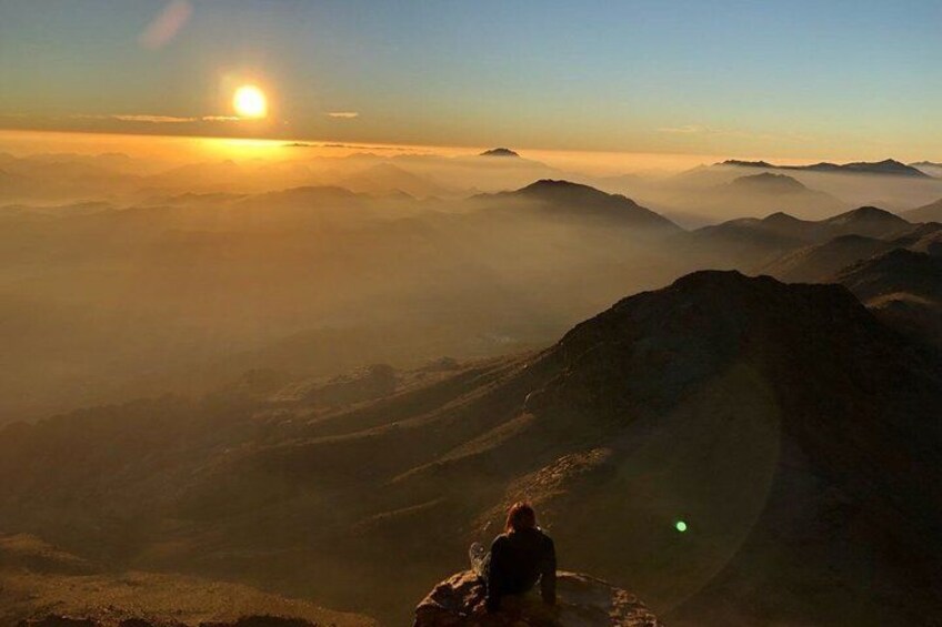 A person sitting on a mountain peak during sunrise with a view of layered mountains and a sun in the background. 5