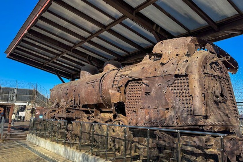A rusted, large, cylindrical locomotive with a covered roof, displayed outdoors with a fence and a building in the background. 4