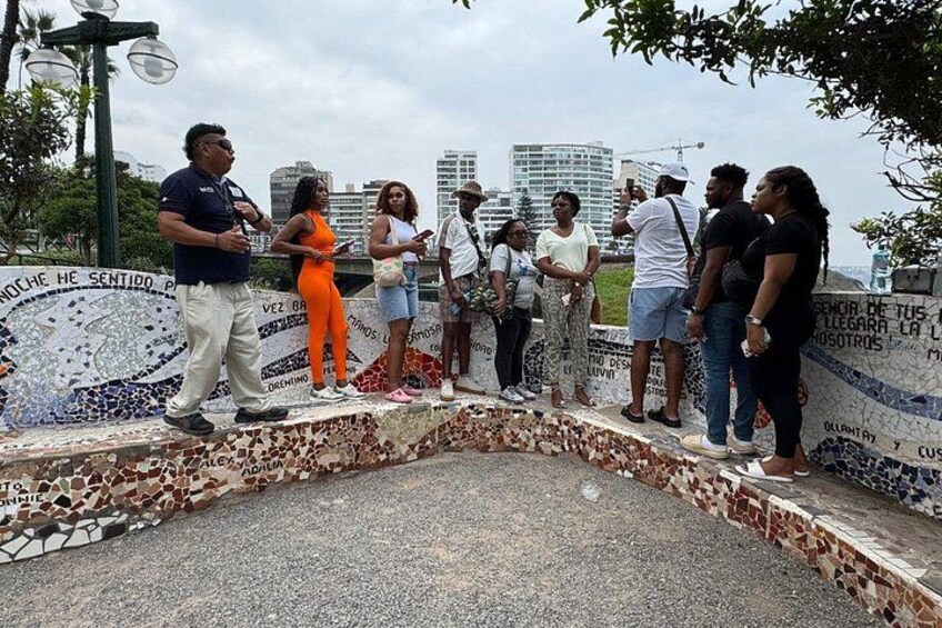 Un grupo de personas sobre una plataforma con baldosas y un muro de mosaicos, con un paisaje urbano al fondo y un farol. 2