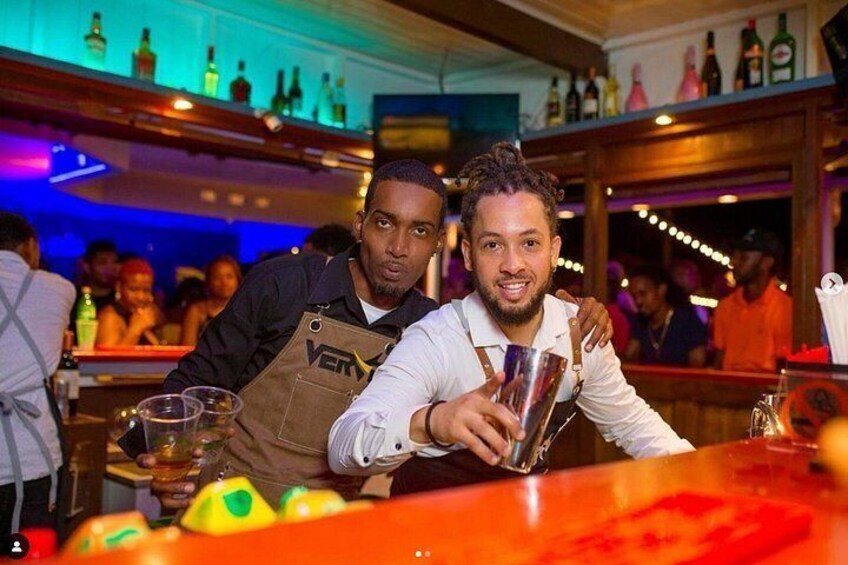 Two bartenders at a bar with bottles and glasses on the shelves. 5