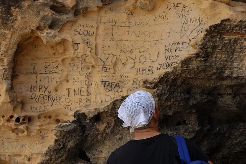 Una persona con un pañuelo blanco en la cabeza frente a un muro de piedra con varios nombres y mensajes escritos. 3