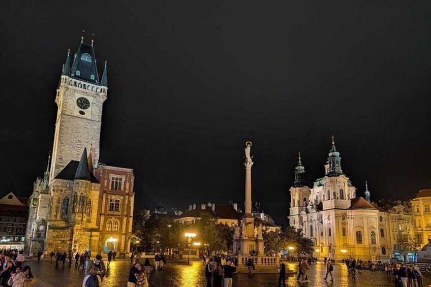 A historic square with a prominent clock tower, a tall monument, and ornate buildings under night lighting. 4