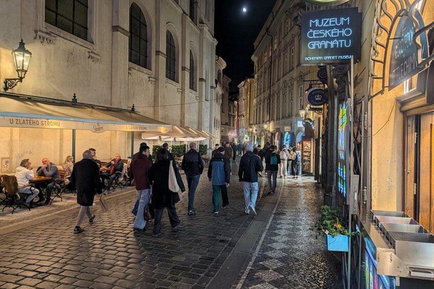 A cobblestone street at night with people walking, cafes, and a museum sign. 2