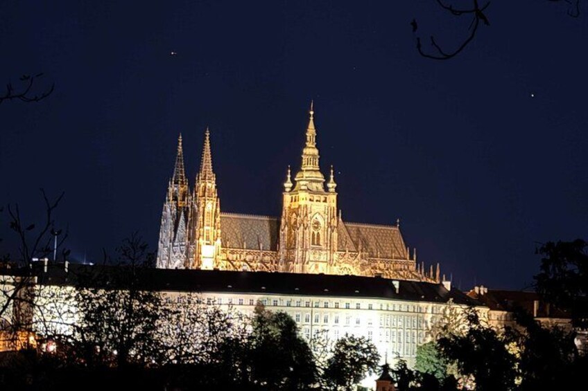 A large illuminated building with multiple spires and a prominent central tower, surrounded by trees. 2