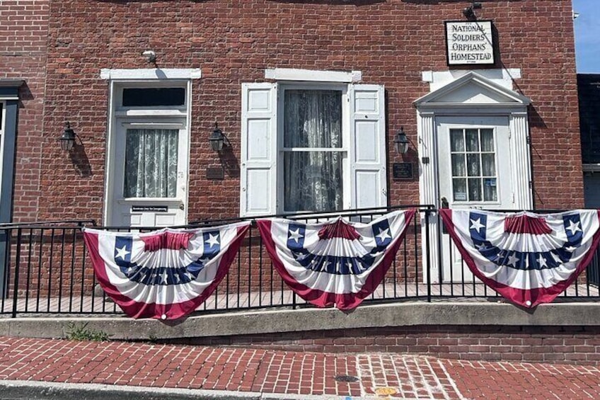 A brick building with white shutters, a sign reading "National Soldiers' Orphans Homestead," and American flags draped over the fence. 1