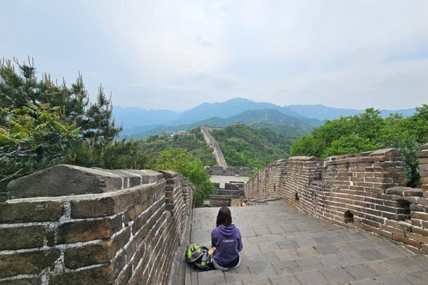 A person sitting on a stone platform overlooking the Great Wall of China with mountains in the background. 1