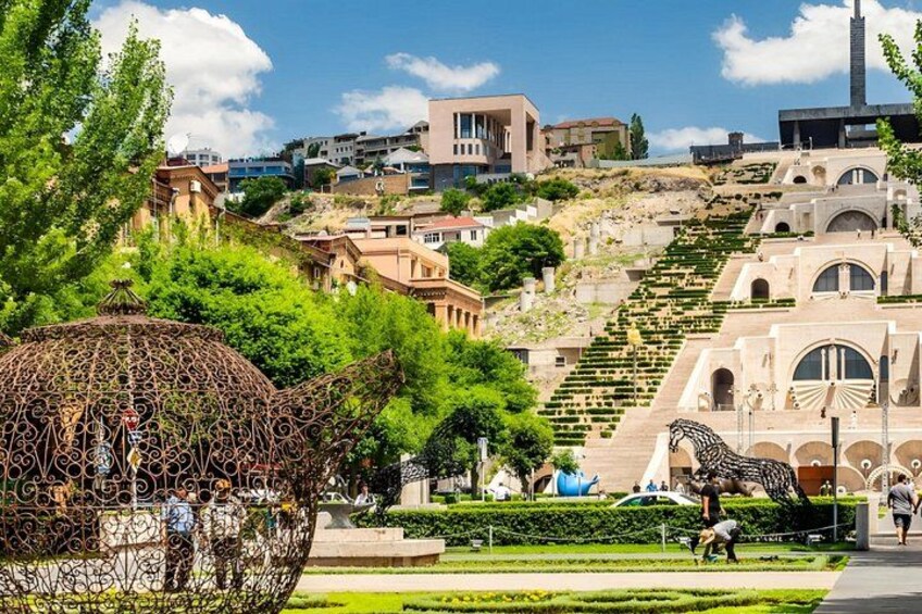 A park with a large metal sculpture, a staircase, and a building with arches. 3