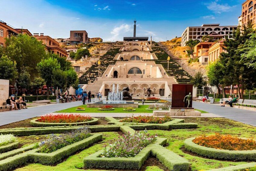 A public square with a central fountain, surrounded by geometrically shaped flower beds and a large building with a dome and steps in the background. 2