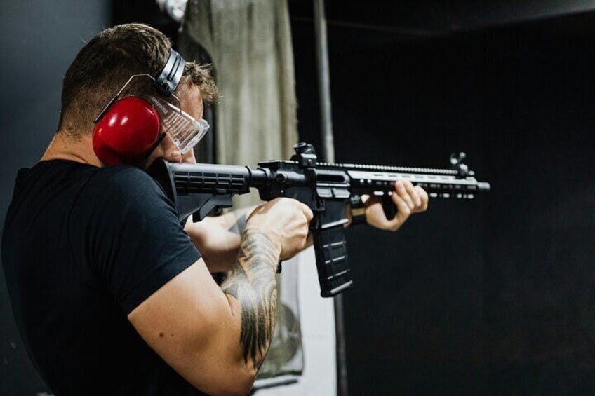 A person aiming a rifle with a scope, wearing red earmuffs, and holding a magazine. 1