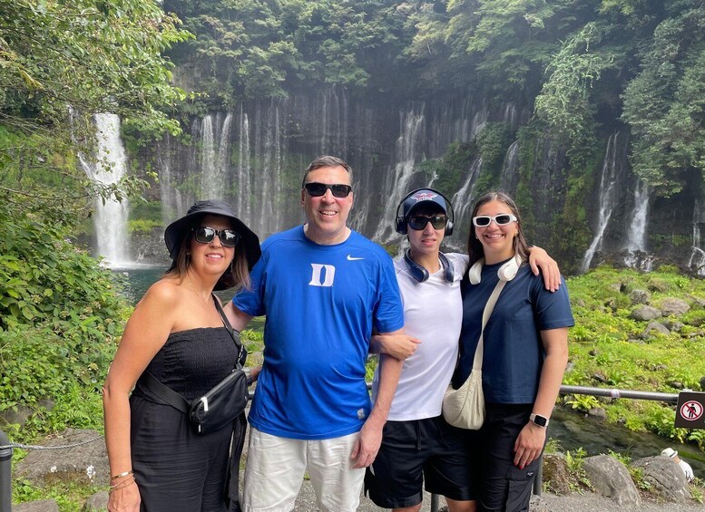 A group of people standing in front of a waterfall with lush greenery. 1