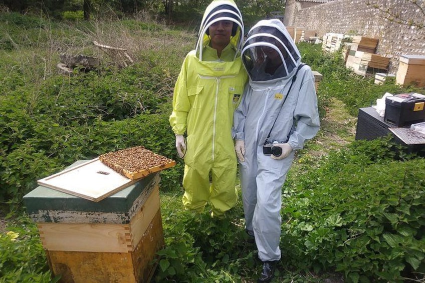 Two people in protective suits standing next to a beehive in a green, leafy outdoor setting. 1