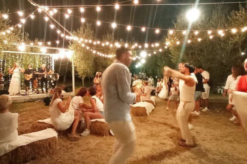 An outdoor event with string lights, a stage with musicians, and people sitting on hay bales. 2