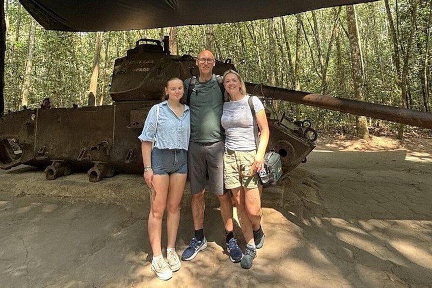 A group of people standing in front of a large, rusted military tank in a forested area. 3
