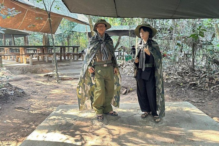 Two people in camouflage attire standing under a canopy in a forested area with picnic tables in the background. 2