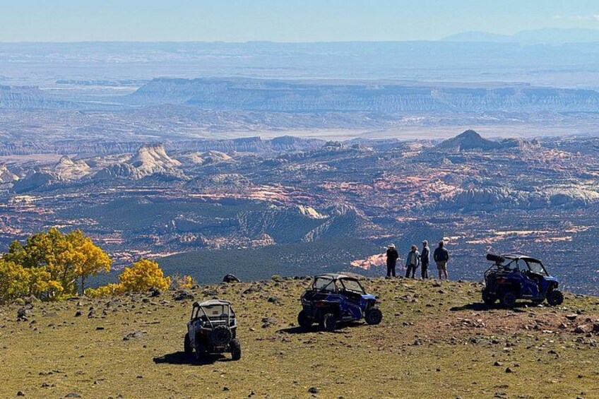 A group of people standing on a hill overlooking a vast landscape with mountains and a valley. There are two off-road vehicles parked on the ground. 4