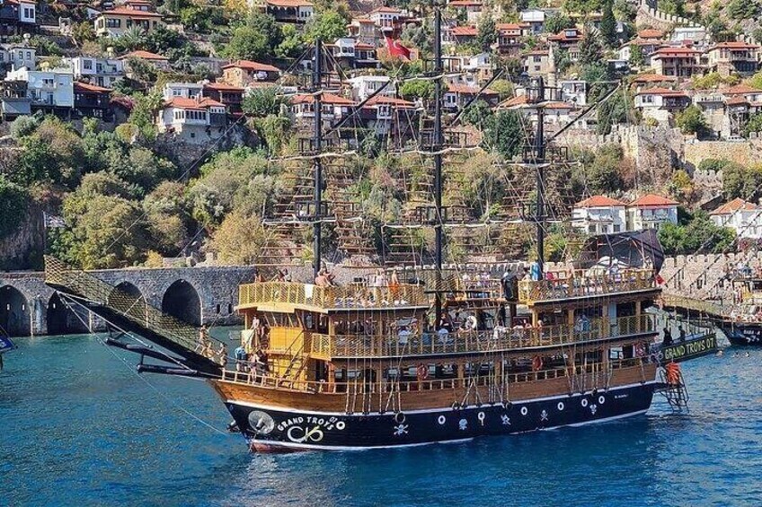 A tall ship with multiple decks and a large mast, docked near a stone bridge and residential buildings on a hillside. 1