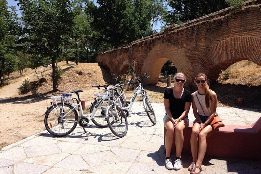Two people sitting on a bench with bicycles parked nearby and a stone bridge in the background. 1