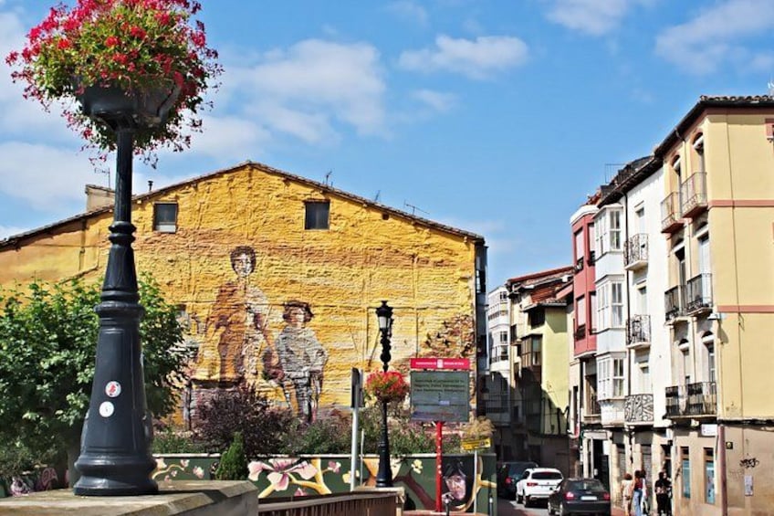 A street with a mural on a building, a street lamp, and a flower basket. 3