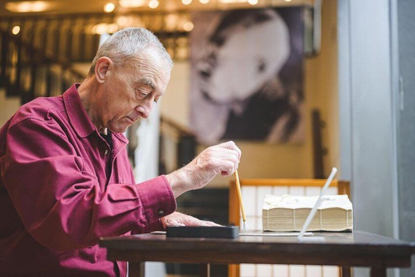 An elderly person in a red shirt sitting at a table with a book and a pen. 1