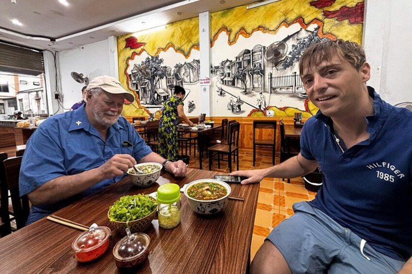 Two people sitting at a wooden table with bowls of food, a mural on the wall, and a plant in the background. 2