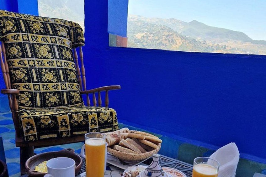 A blue-painted room with a patterned chair, a table with food and drinks, and a view of mountains. 3