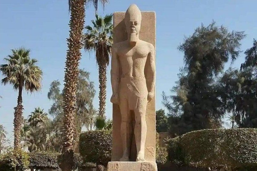 Eine große Steinstatue einer Figur mit Kopfschmuck, die auf einem Sockel steht, umgeben von Palmen und anderer Vegetation. 5