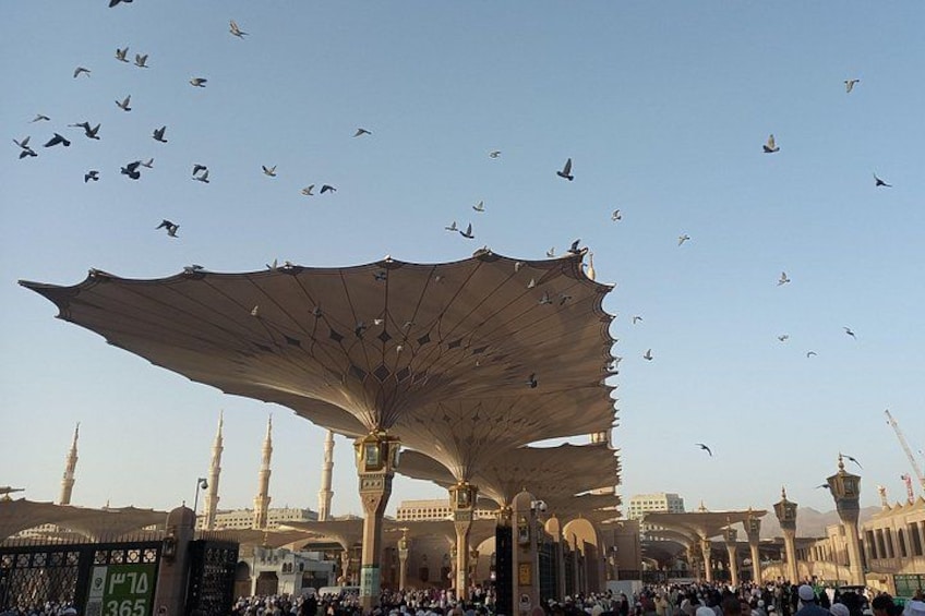 A large open structure with a canopy, surrounded by tall buildings with pointed tops, and a crowd of people below. 1
